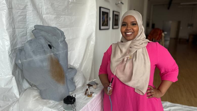 A young woman in a pink hijab and dark pink dress applies paint to a sculpture of two hands clasping.