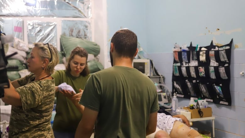 Three medics in fatigues gather around a man in a hospital gown on a stretcher in a room filled with medical gear. 