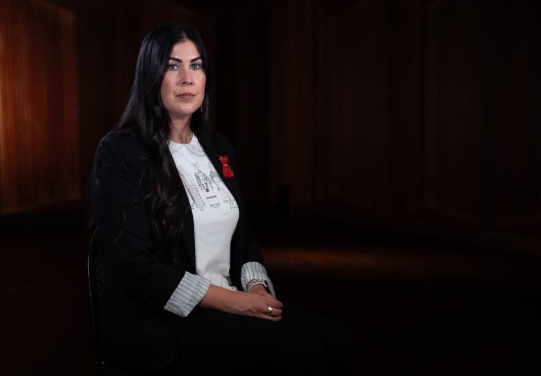 A woman with long black hair and dark eyebrows, with a red dress pin on a black blazer, sits in a darkened room, staring directly at the camera.