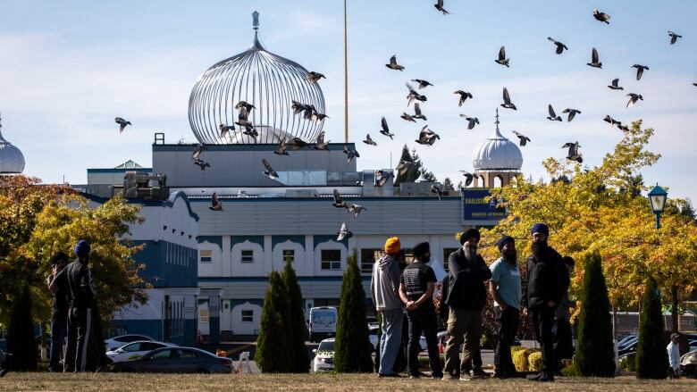 Sikh men stand in the shadow of a large gurdwara as birds fly above them.