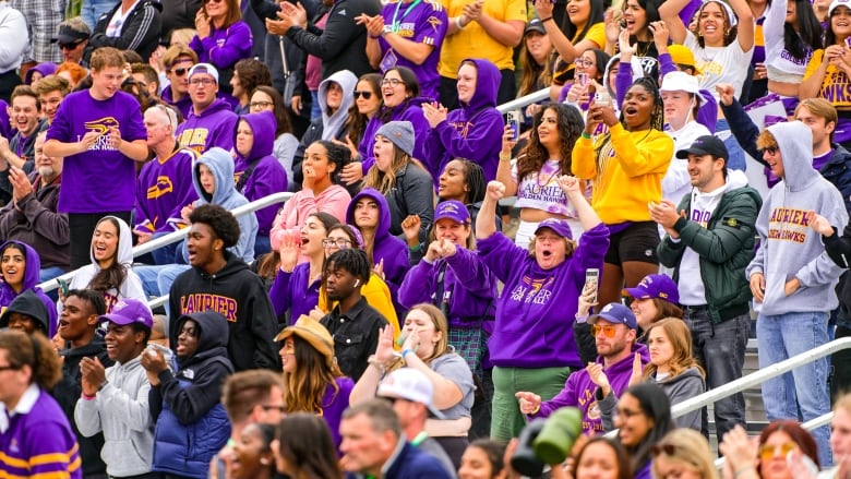 A large group of fans wearing yellow and purple Laurier colours cheer in the stands.