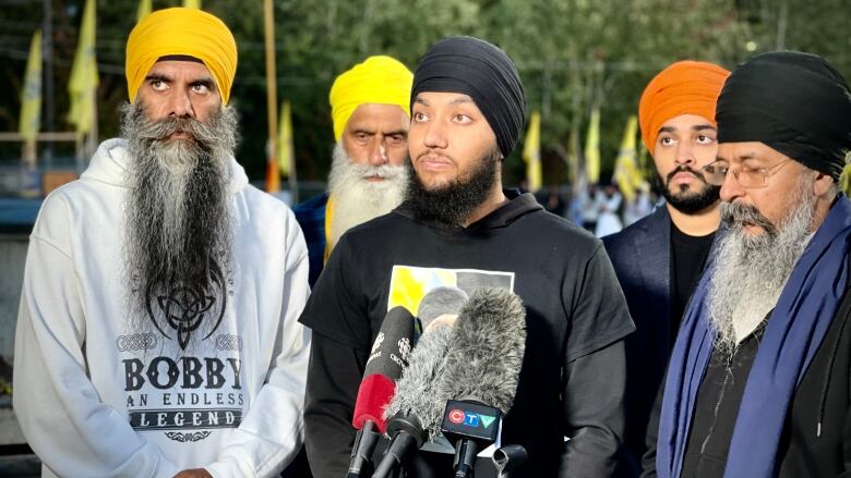 A Sikh man speaks to a collection of mics, flanked by other Sikh men.