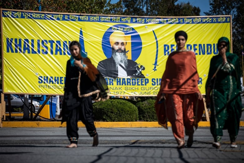 Three women walk past a yellow flag with a Sikh man, with words about a Khalistan referendum.