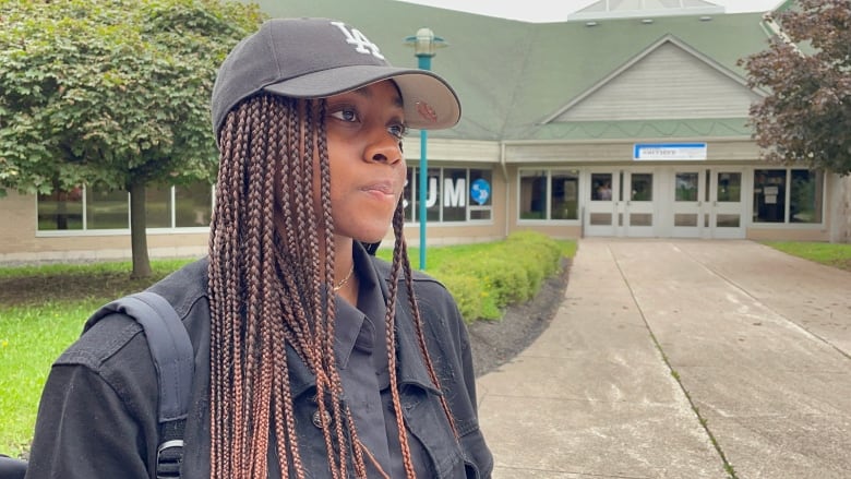 A black girl with brown coloured braided hair. Wearing a black cap, black shirt and a backpack. 