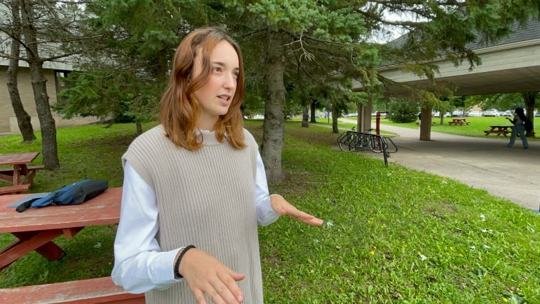 A white girl with brown hair, speaking while moving her hands. She is wearing a light blue shirt and a light grey sweater.