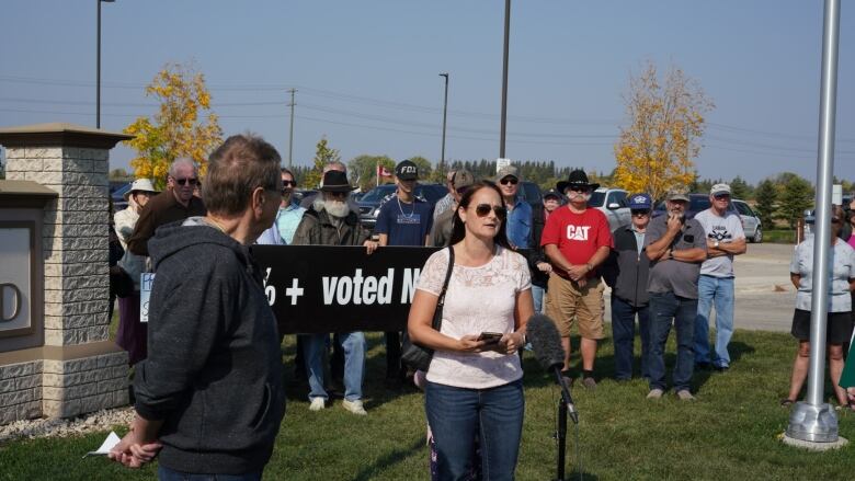 A woman with shoulder-length dark hair and sunglasses speaks to a journalist outside a council building.