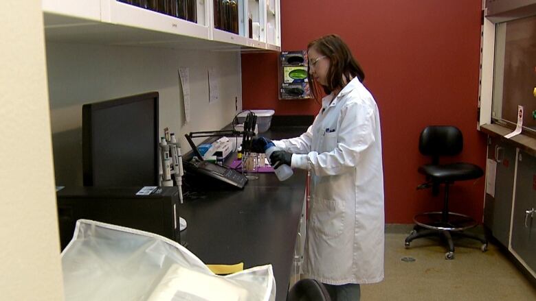 A woman in a white lab coat stands at a counter with beakers on it. 