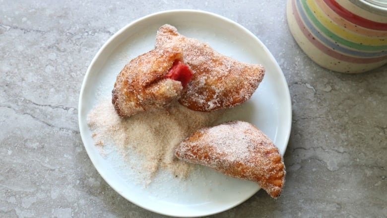 Three half moon pockets sit on a plate. One is half eaten and red can be seen inside. They are topped with sugar. 