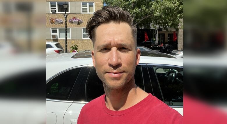 A head-and-shoulders image of a man in a red shirt, standing outside in the sun, a street in the background.