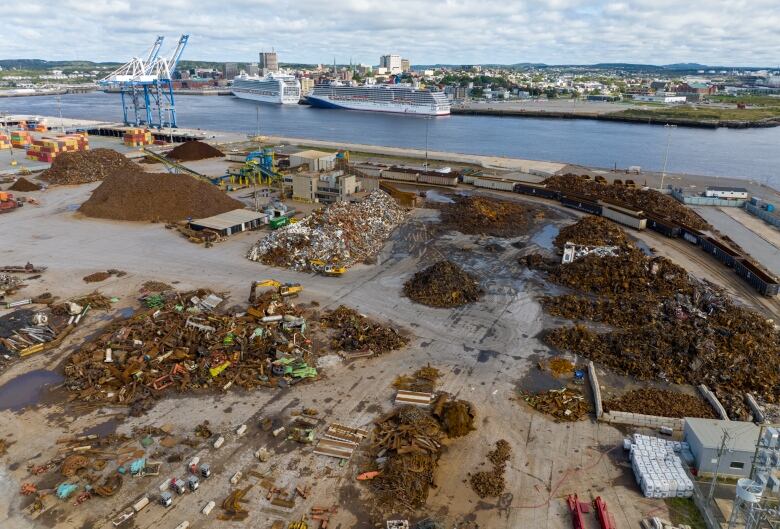 Several piles of scrap material sit on an industrial dock with two large cruise ships moored across the harbour. 