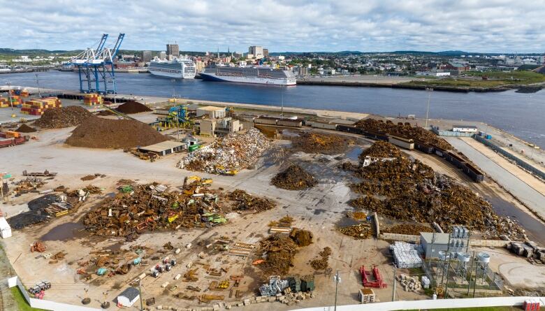 Several piles of scrap material sit on an industrial dock with two large cruise ships moored across the harbour. 