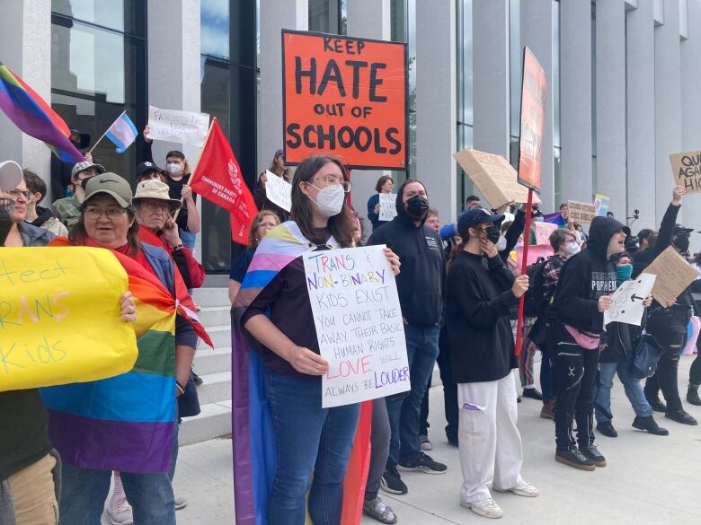 A crowd of people with rainbow flags and signs. A big orange sign says 