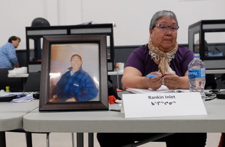 A woman sits behind a table inside a room, beside a framed photograph of a man.