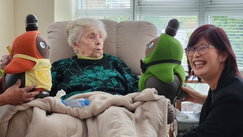 An elderly woman with white hair sits in a reclining arm chair wearing a black sweater with green flowers and a beige blanket over her legs. She is looking at what looks like a plush toy about a foot tall with a surprised and delighted look on her face.