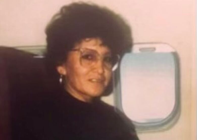 A woman with glasses and short curly hair is wearing a black sweater while pictured sitting next to an oval window on what appears to be an aircraft.