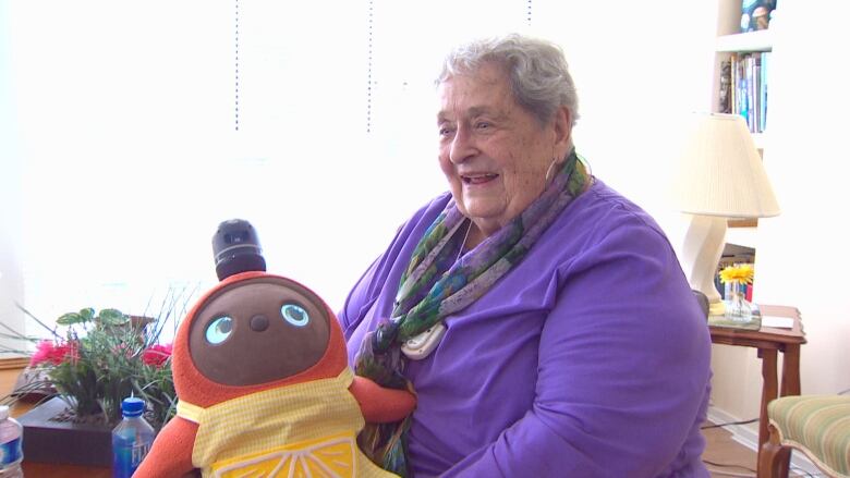 A woman with grey short hair sits holding what appears to be an orange stuffed animal wearing a yellow apron. The woman wears a purple blazer and colourful scarf and a smile.