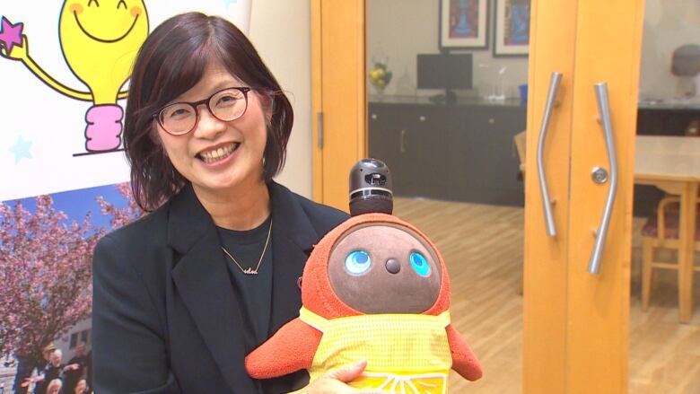 A young Asian woman wearing a black blazer holds an orange plush toy wearing a yellow apron.