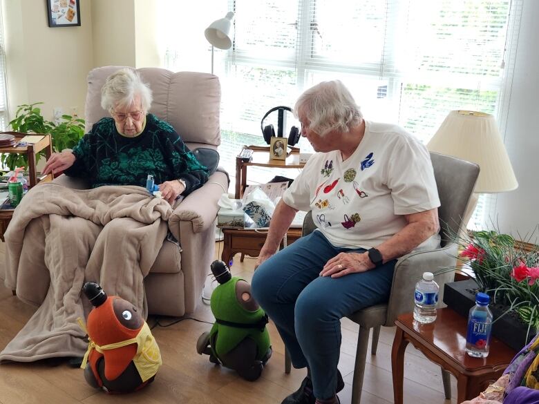 Two elderly women, one in a bed and the other sitting next to her in a chair, look at two robots that resemble plush toys moving on the floor.