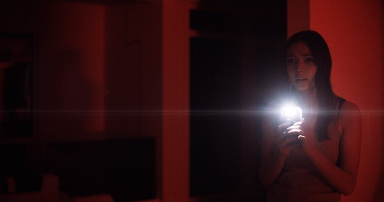 A woman holds a phone with a flashlight emitting from it. The lighting in the room is red, which is making the walls and the woman appear red as well. 