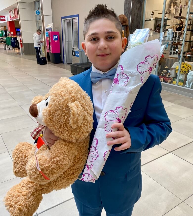 Boy holds teddy bear and flowers at airport.