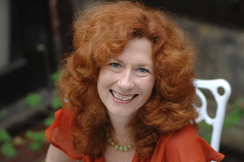 A woman with long, wavy red hair is smiling and wearing a crimson top.