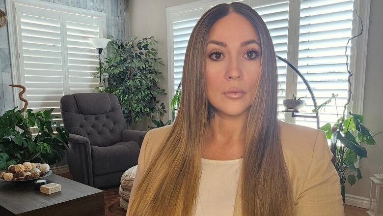 A woman with long hair sits in a living room looking at the camera.