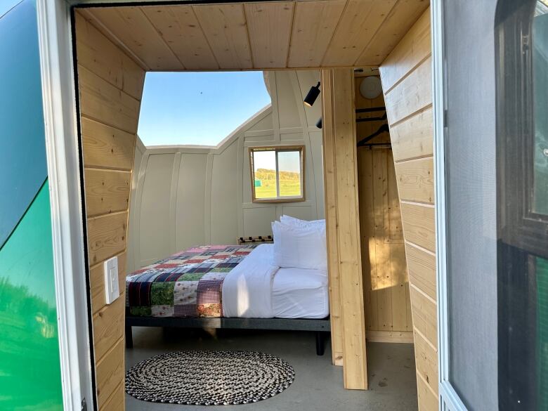 An open door to a bed with a handmade quilt and oval rug, on a grey concrete floor.