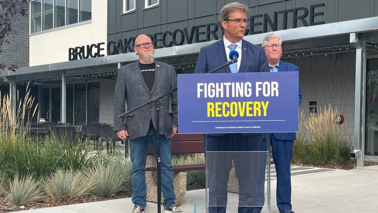 Three men stand behind a podium and in front of a building.