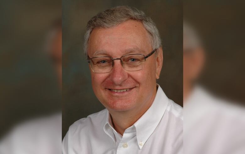 A man with white hair and glasses is seen from the head and shoulders. He smiles at the camera and wears a white collared shirt.