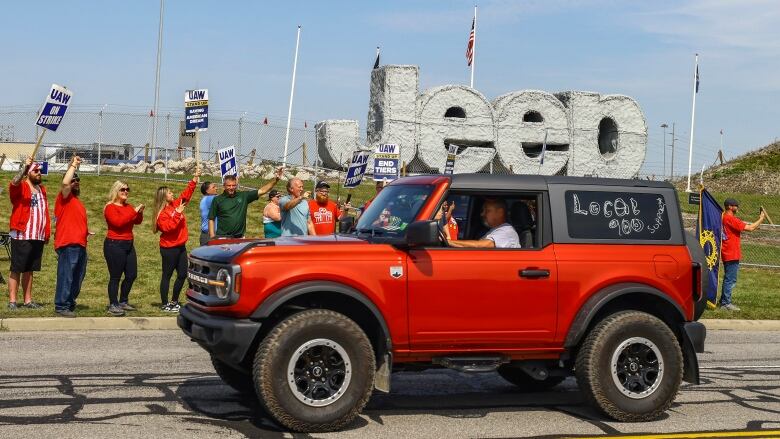 People hold flags while inside a jeep driving by workers picketing at an automotive plant.