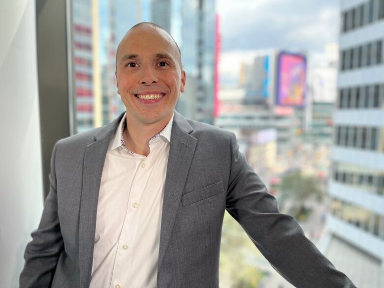 A man with a shaved head wearing a grey suit and white dress shirt stands smiling in front of a office window in downtown Toronto, with tall buildings in the background. 