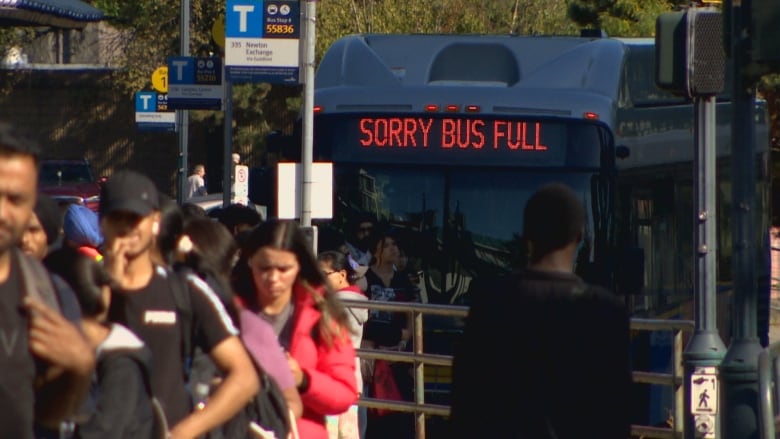 A long line of people wait at the Surrey Central Station with a bus displaying, Sorry, Bus Full.