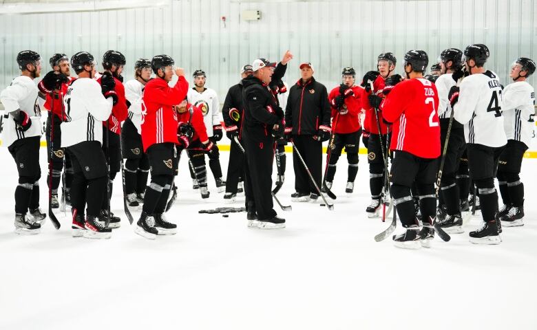 A hockey coach points in the air as he's surrounded by his players during a practice.