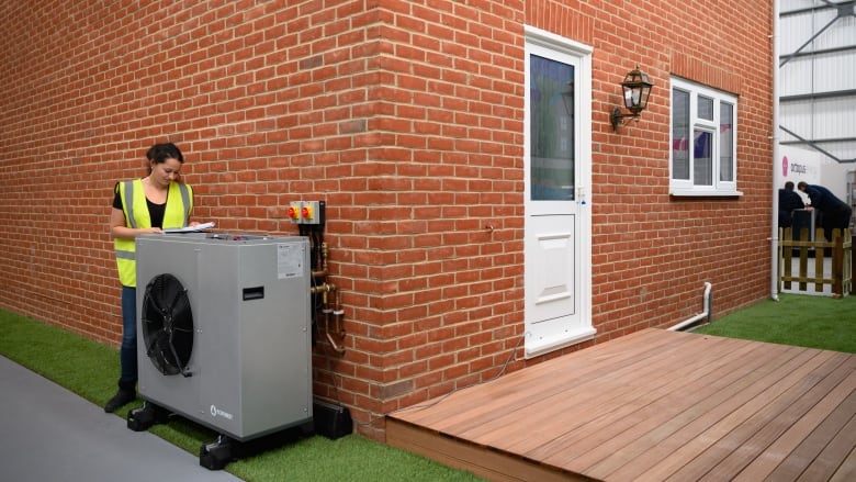 A woman in a fluorescent safety vest writes on a clipboard on the grey outdoor unit of a heat pump at the corner of a brick house.