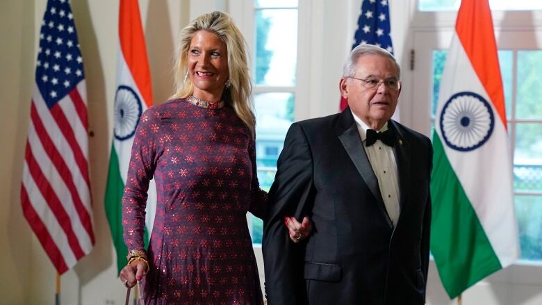 A man and woman are shown with arms locked in formal wear entering a room that contains several flags.