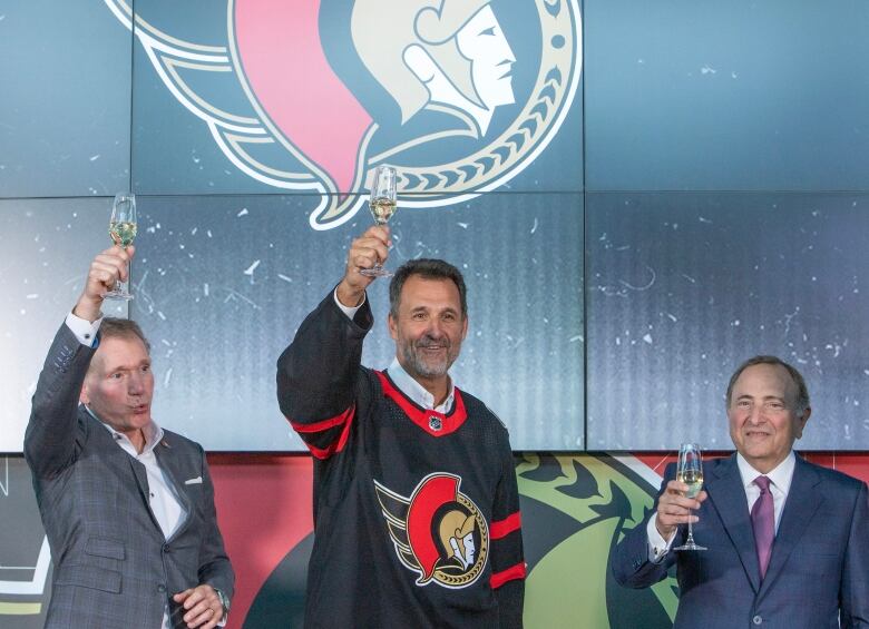 Three men raise champagne glasses in front of a hockey team's logo.