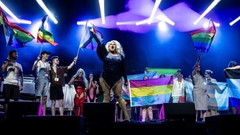 People standing on stage, with a drag performer in the centre, all waving pride flags in the air.