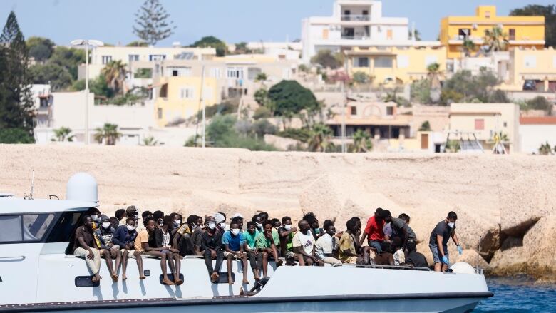 Several people crowded on the deck of boat sailing past buildings in the background.