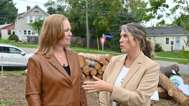 A woman in a brown leather coat with shoulder length hair speaking with a woman with a tan suit jacket with hair in a ponytail.