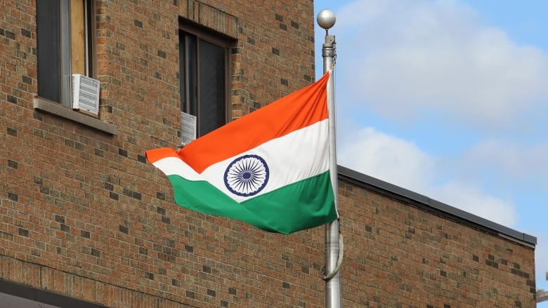 The flag of India waves in the wind on a flag pole in front of a brown brick building.