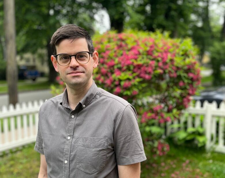 A man wearing glasses and a short-sleeved grey shirt stands outside in front of a flowering bush.