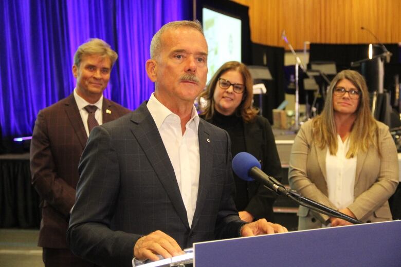 A man speaks at a podium, while flanked by three other people behind him, to his left.