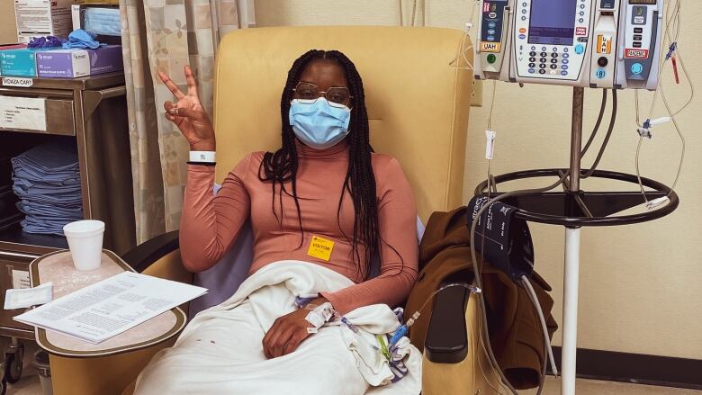 A woman flashes a V for Victory sign while wearing a medical mask and sitting in a chair beside an IV stand and other medical equipment.