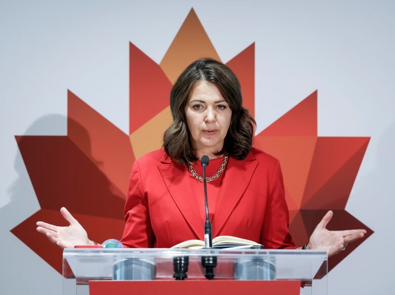 Alberta Premier Danielle Smith speaks to the media in Calgary while standing at a podium in front of a large maple leaf backdrop.