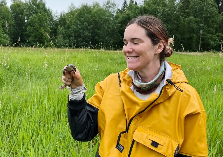 A woman wearing a yellow jacket holds a frog in a field.