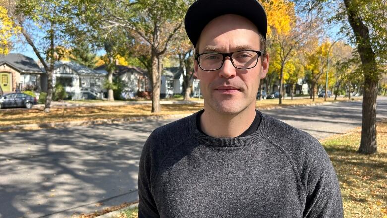 A man can be seen wearing a black sweater with trees behind him, he is wearing a cap and black rimmed reading glasses. 