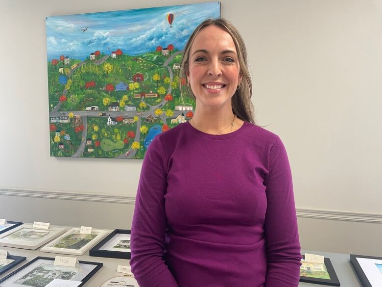 A woman smiles and wears a purple shirt. She stands in front of a colourful and vibrant painting. 