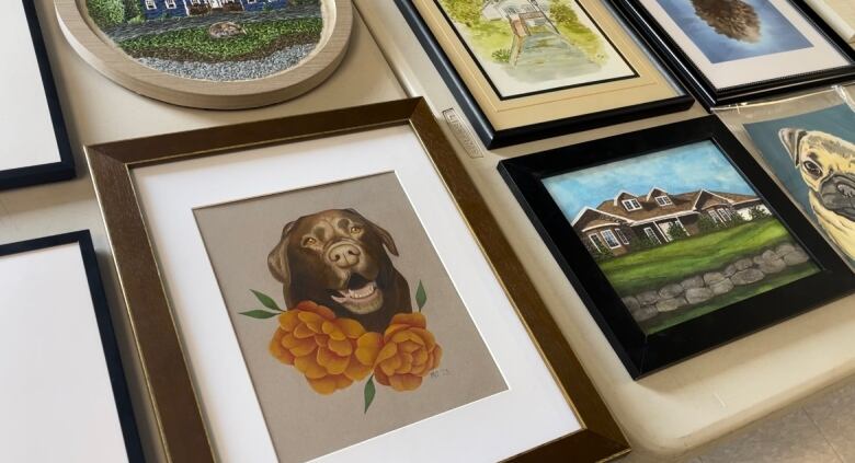 An array of framed drawings and paintings are laid out on a table. The most prominent is a chocolate lab with orange flowers underneath it.