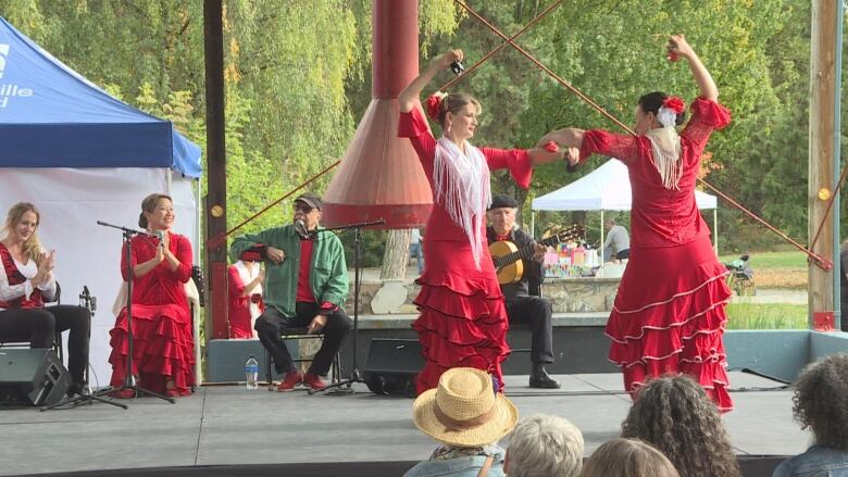 Two flamenco dancers hold up their arms wearing red dresses while an audience listens.