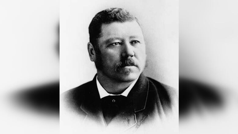 Black and white photo of the head and shoulders of a man with short wavy hair and a moustache. He is looking off to the right of the frame.